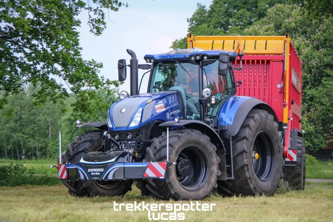 Blue tractor with front weight bumper and large rear trailer on grass, trees in background, text overlay at bottom