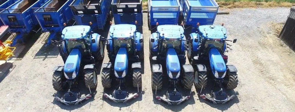 Four blue tractors fitted with front weight bumpers are parked in a row facing blue trailers on a dirt lot