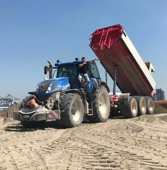 Blue tractor with a front weight bumper attached, pulling a raised red and white dump trailer on sandy ground