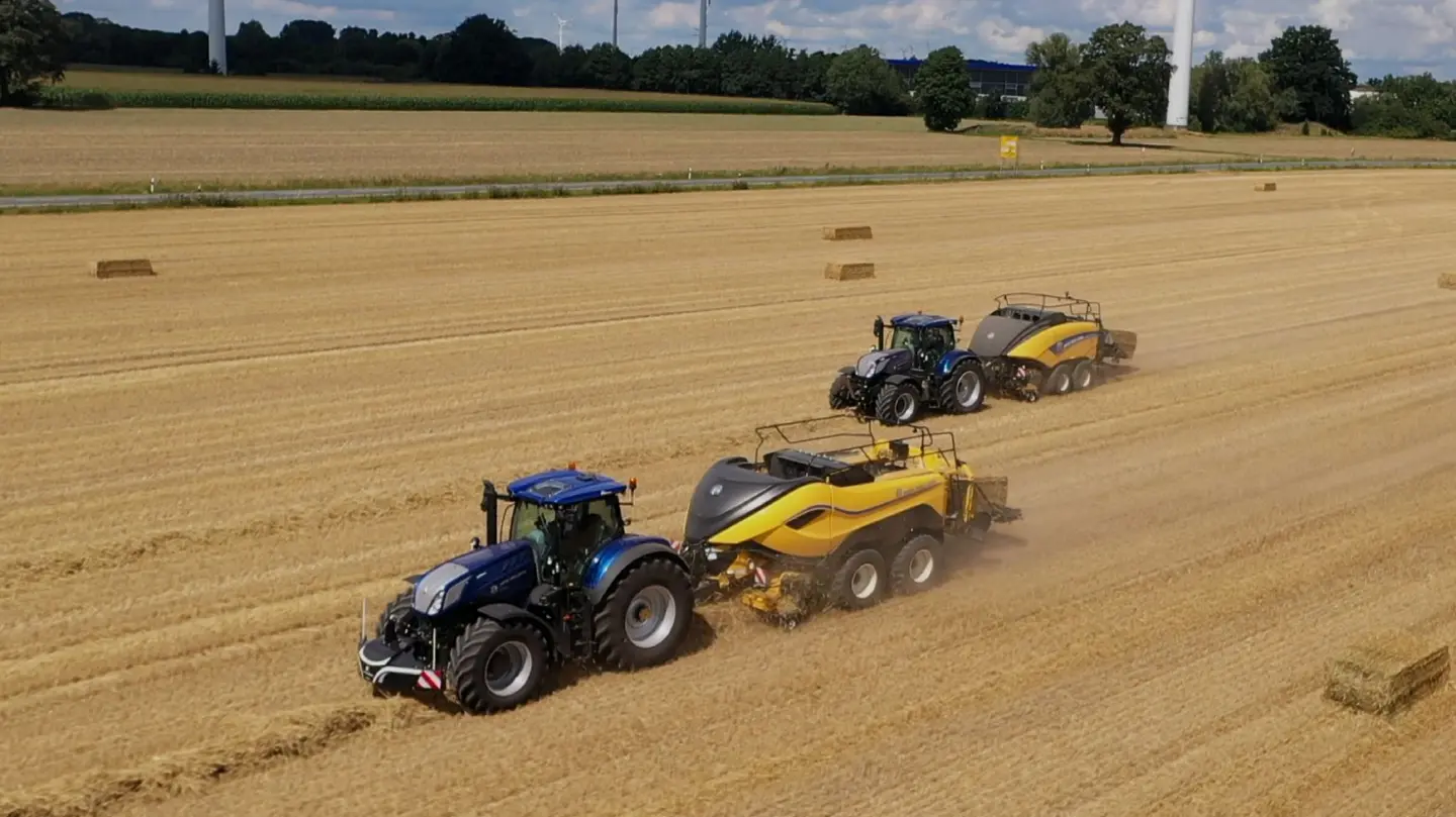 Two blue tractors with front weight bumpers pull yellow balers through a harvested field with hay bales scattered around