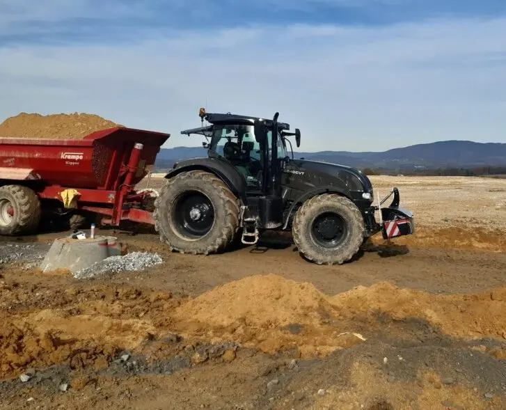 Black tractor with a front weight bumper and large tires pulling a red dirt trailer on a construction site