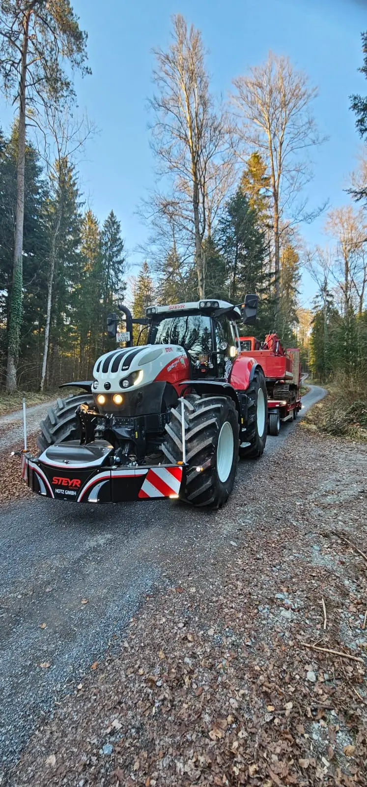 A tractor with a front weight bumper labeled STEYR HOLZ GMBH is parked on a forest road with a red trailer.