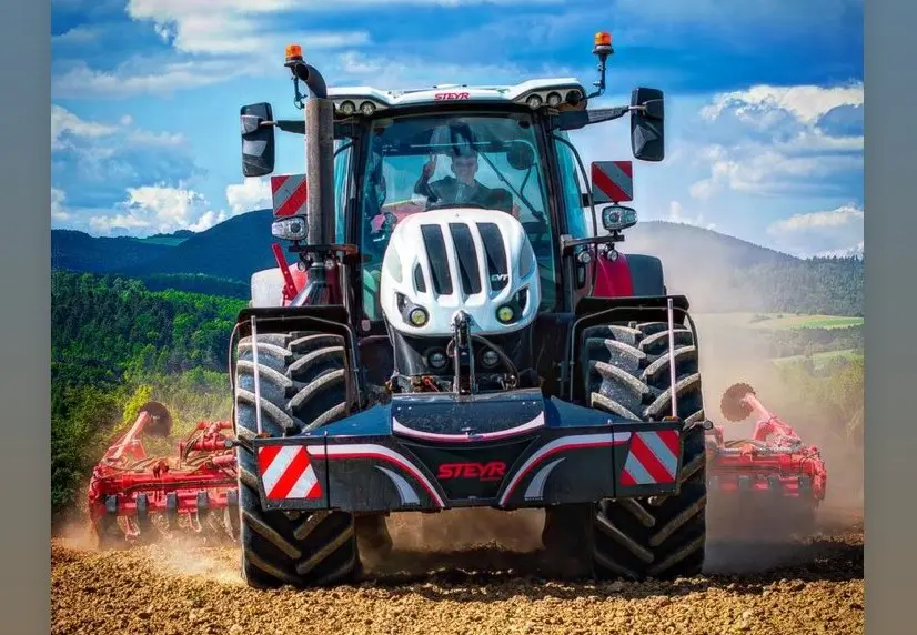 A tractor with a Steyr front weight bumper is plowing a field on a sunny day with hills in the background