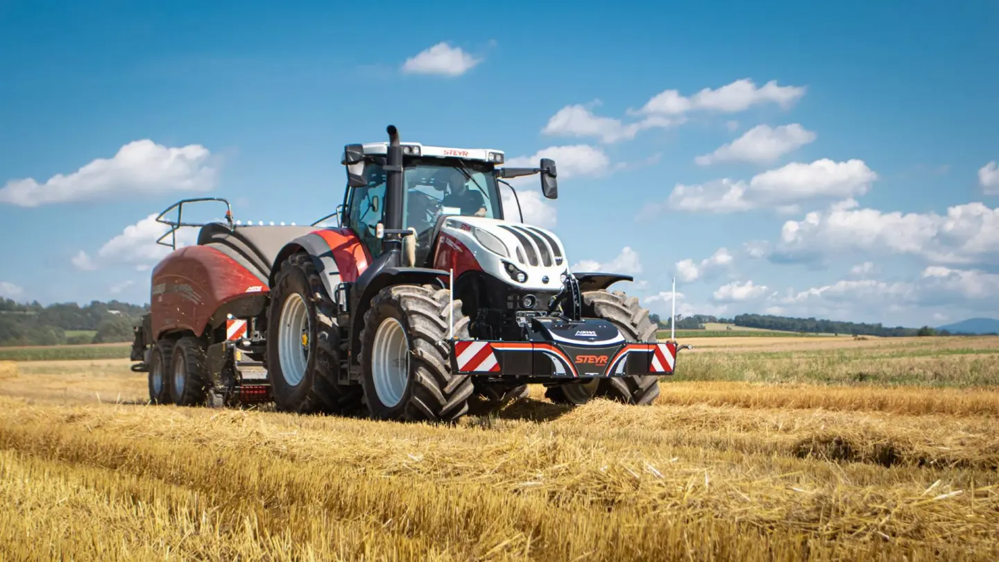 A tractor with a Steyr front weight bumper attached works in a harvested field under a blue sky with clouds