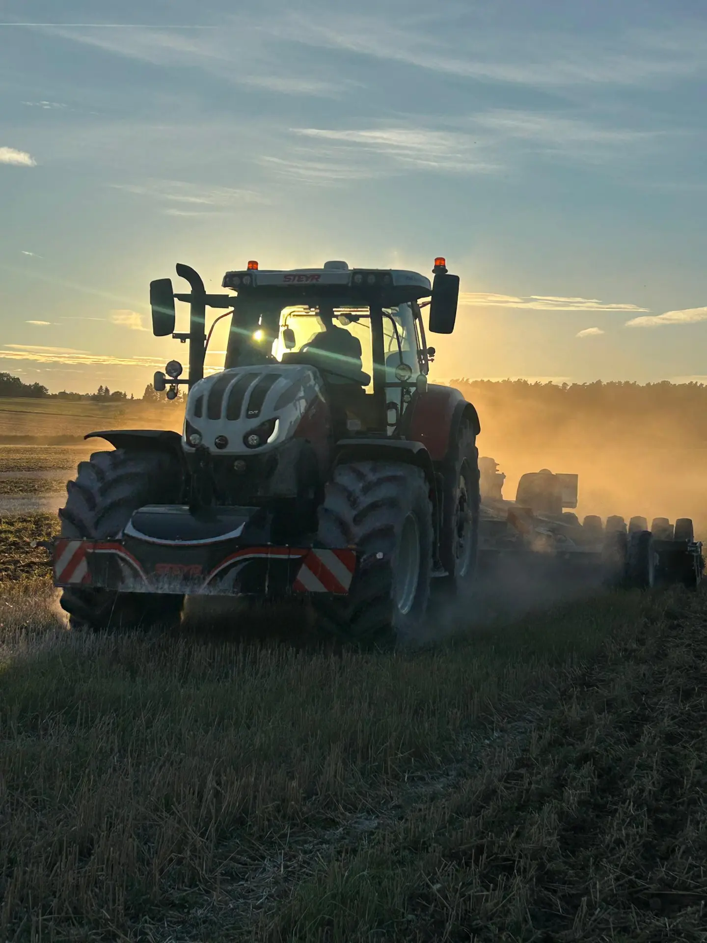 Tractor with a striped front weight bumper working in a field at sunset, kicking up dust as it moves forward