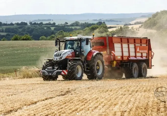 Red and white tractor with a front weight bumper towing a large red trailer through a harvested field