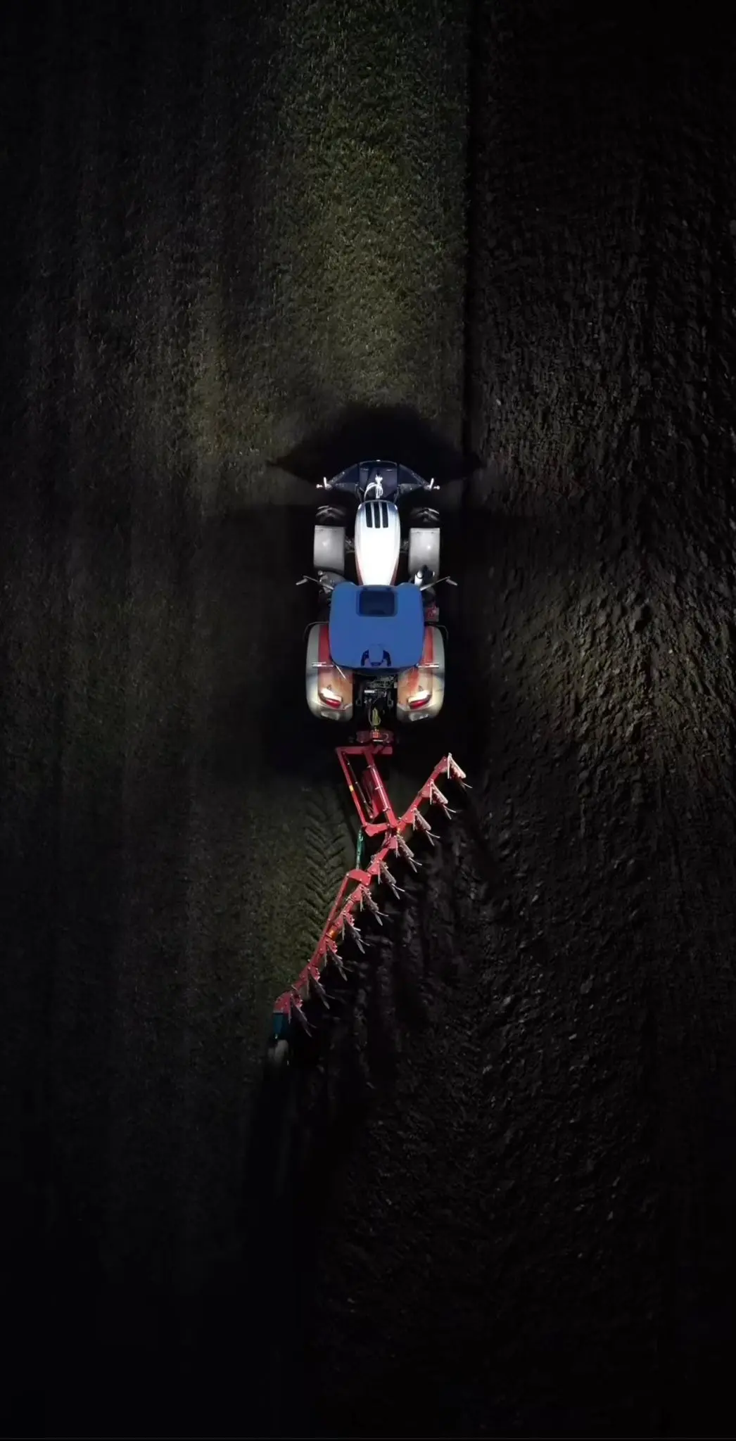 Tractor with a front weight bumper and mounted plough working on a field at night, viewed from above
