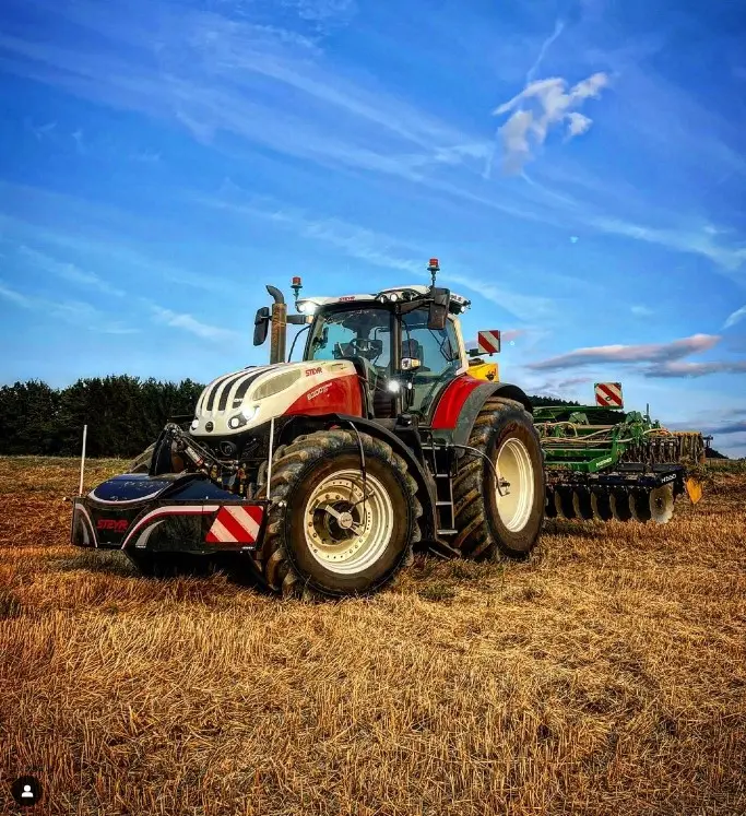 A tractor with a front weight bumper is parked in a harvested field with blue sky and clouds in the background