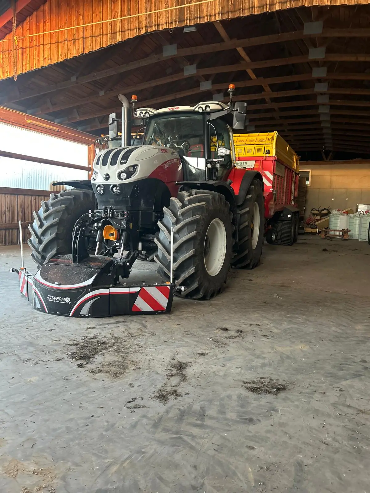 Tractor with a front weight bumper attached, parked indoors and hitched to a yellow and red trailer