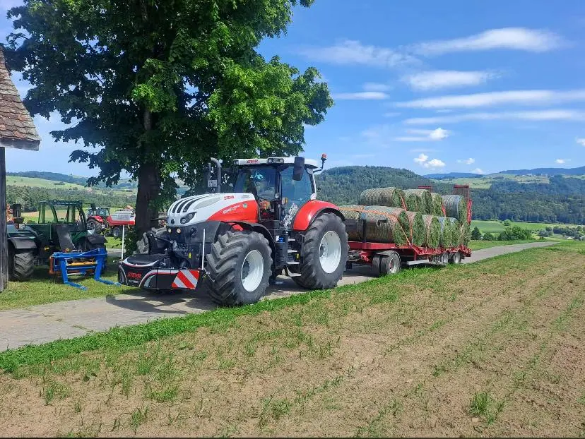 A red and white tractor with a front weight bumper is pulling a trailer loaded with round hay bales on a rural road