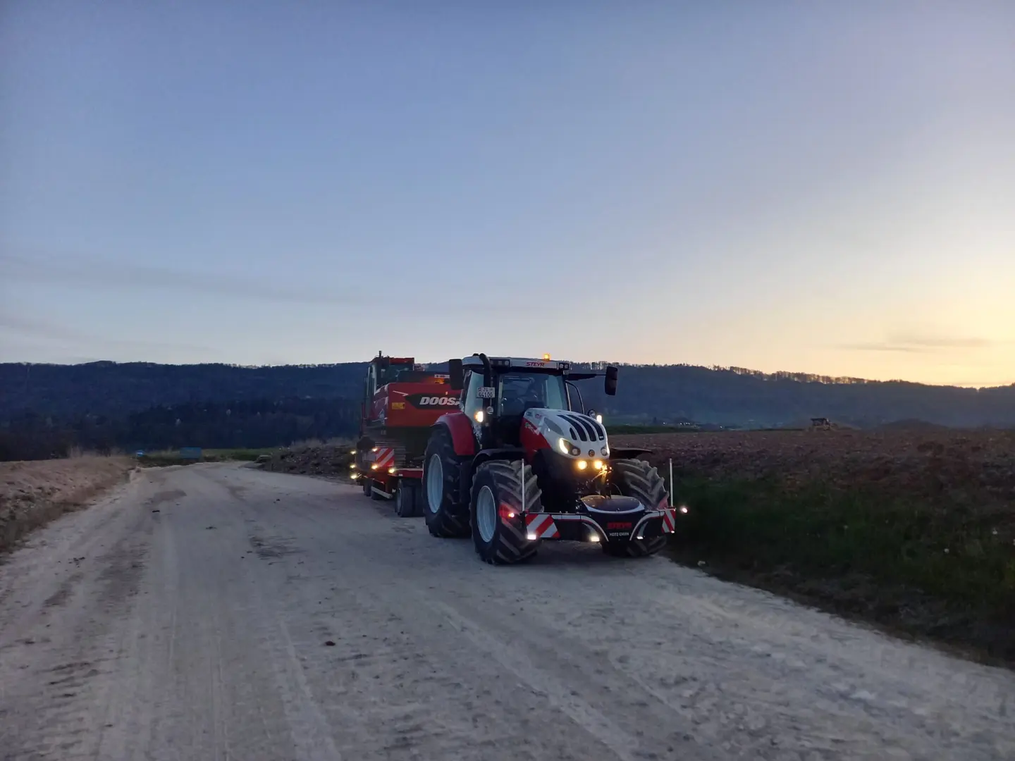 A tractor with headlights on is towing equipment on a dirt road at dusk, fitted with a front bumper marked Steyr.