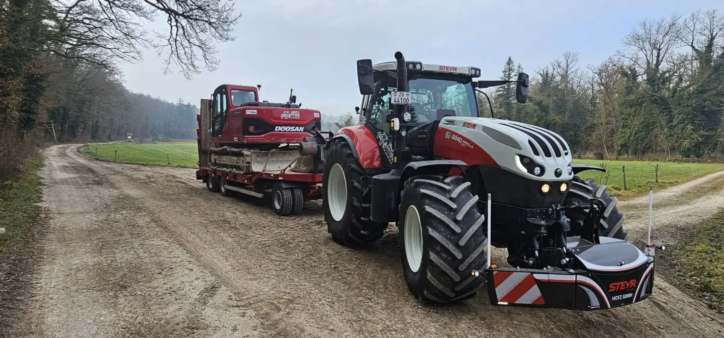 A Steyr tractor with a front weight bumper attached is towing a trailer carrying a red excavator on a dirt road