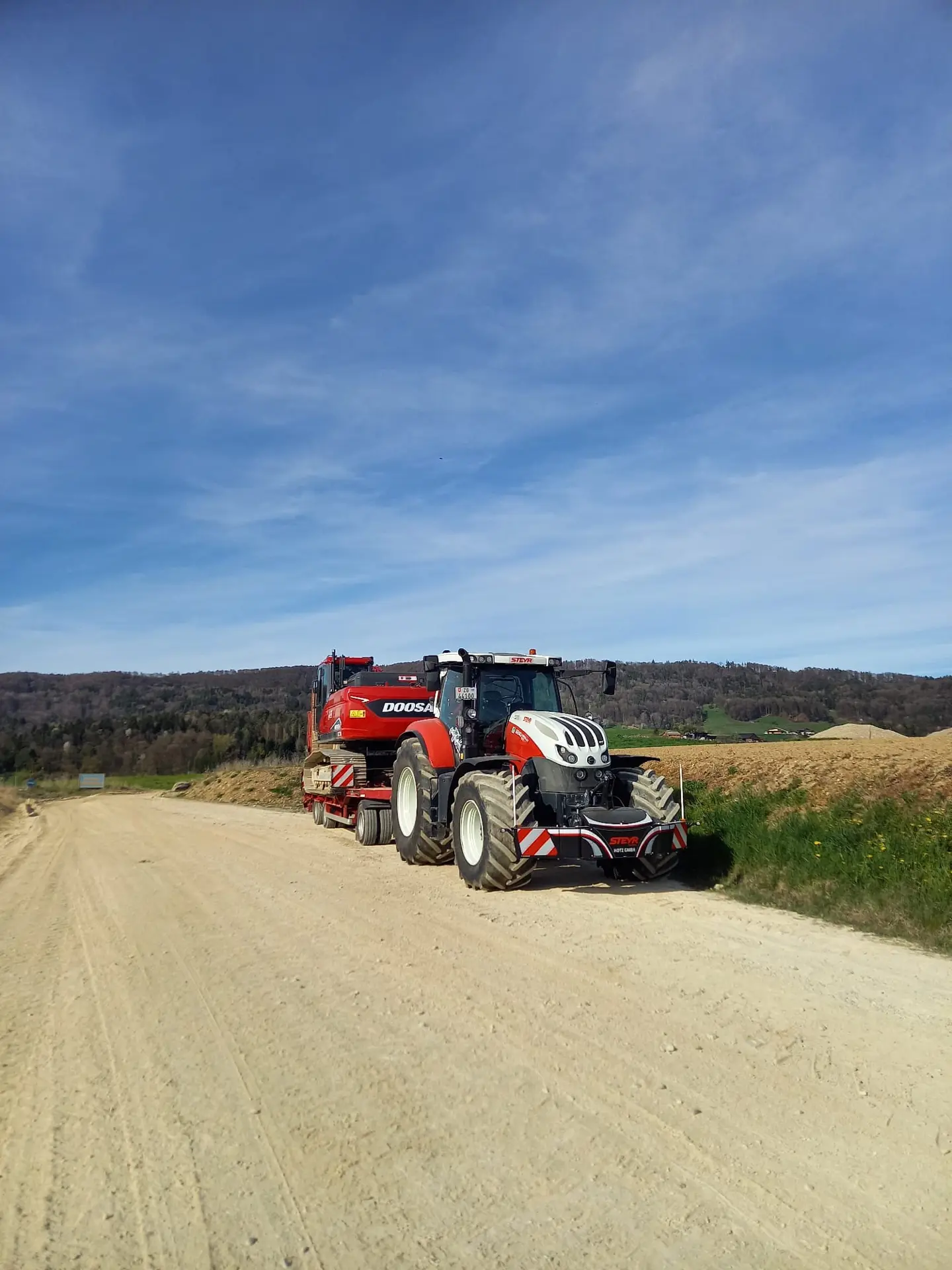 Red and white tractor with a front weight bumper and trailer parked on a dirt road beside grassy fields