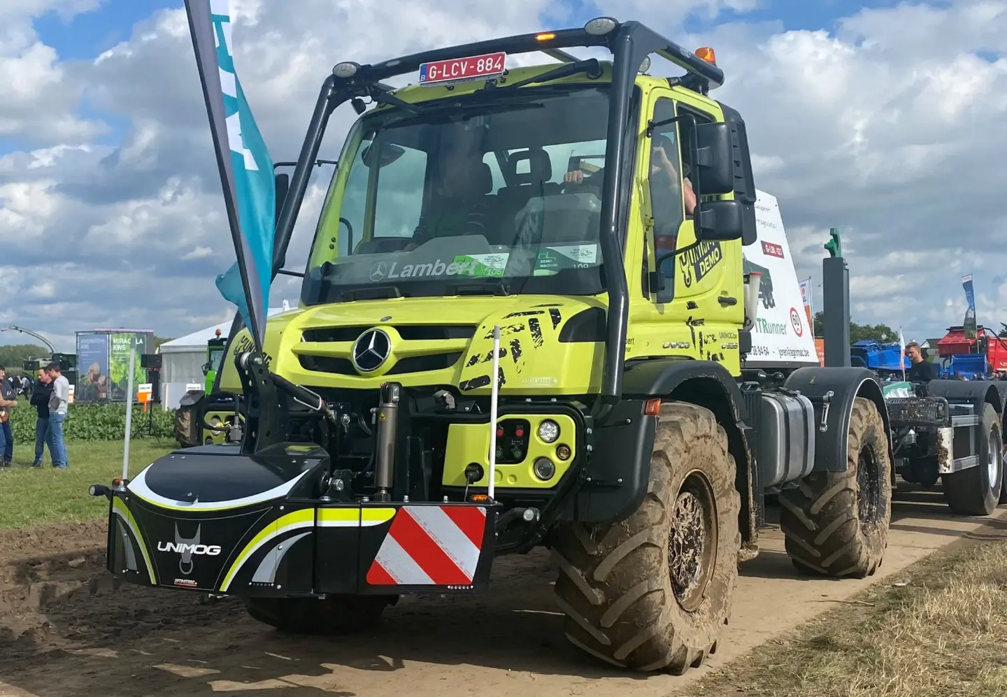 A green Unimog tractor with a large front weight bumper featuring red and white reflective stripes on display outdoors