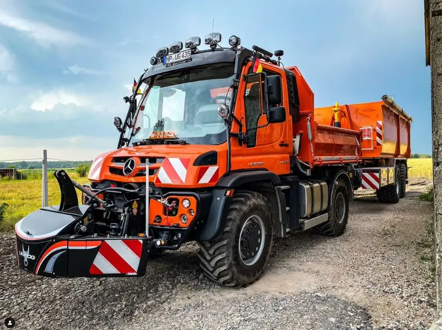 An orange utility vehicle with large tires and a front weight bumper, towing a matching orange trailer on a gravel path