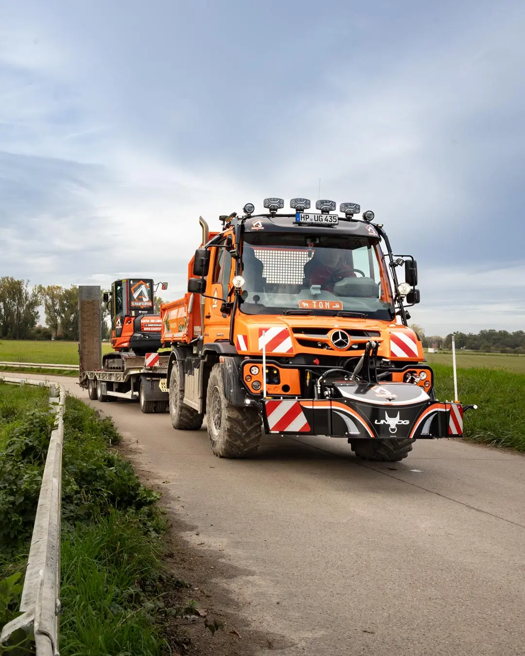 Orange utility vehicle with large front weight bumper and reflective stripes, towing a trailer on a rural road