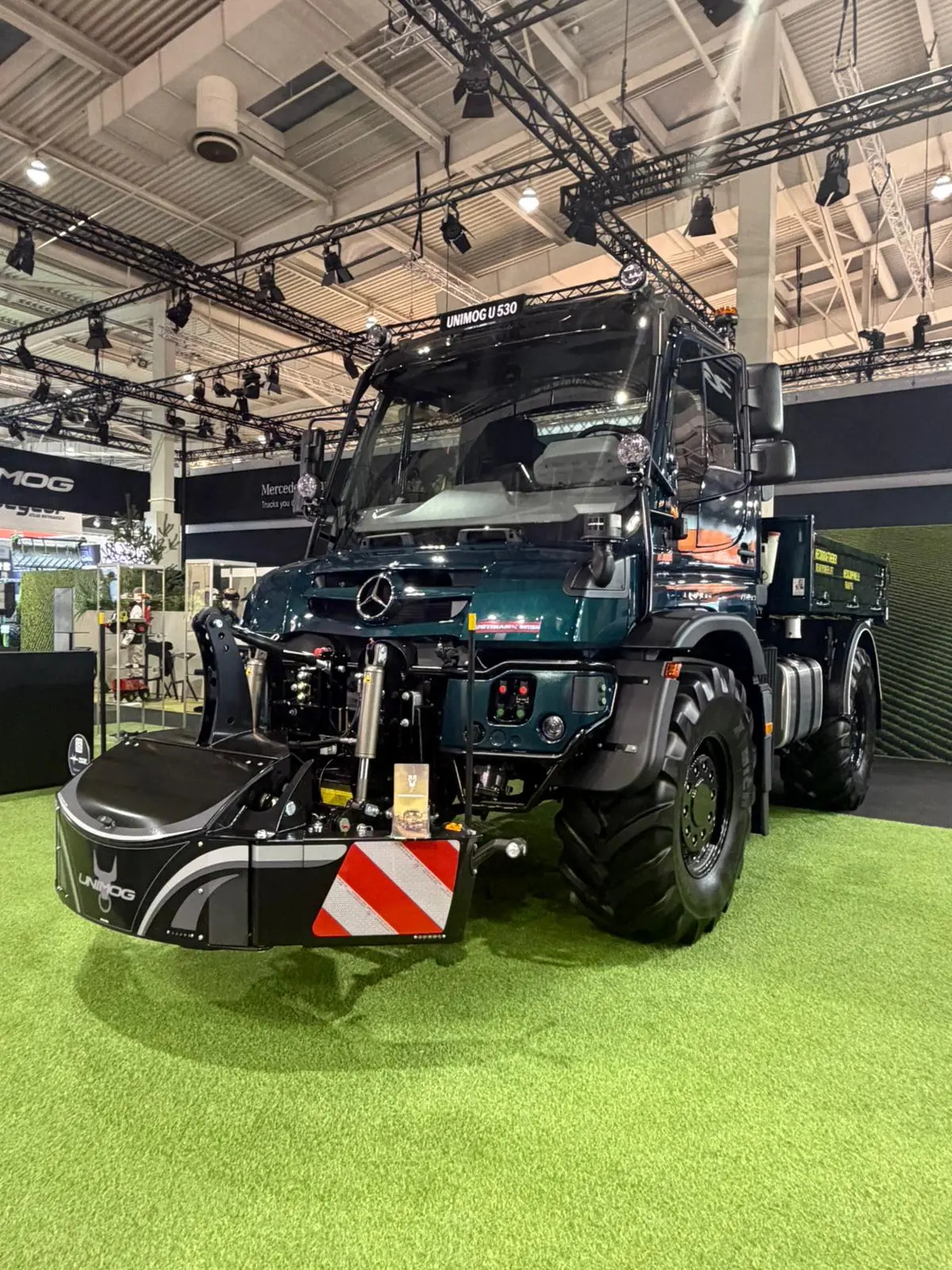 A large dark green utility vehicle with a front-mounted weight bumper featuring red and white reflective stripes