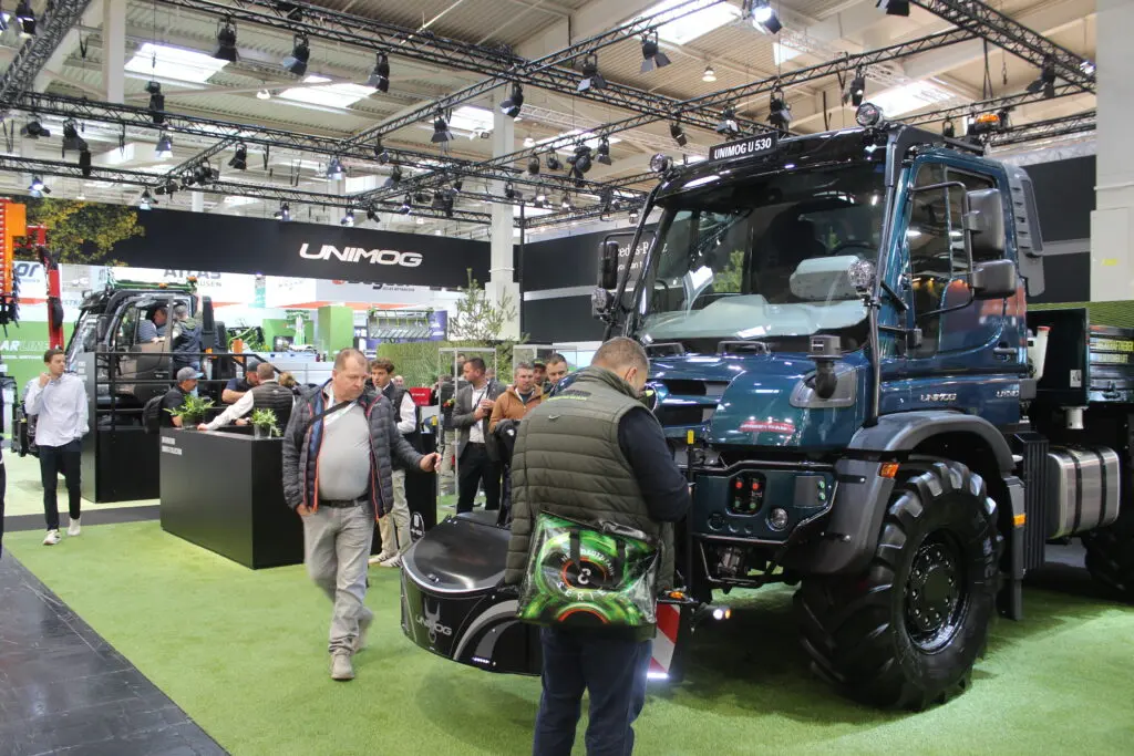 People examine a Unimog vehicle with a front weight bumper attachment at an indoor exhibition booth