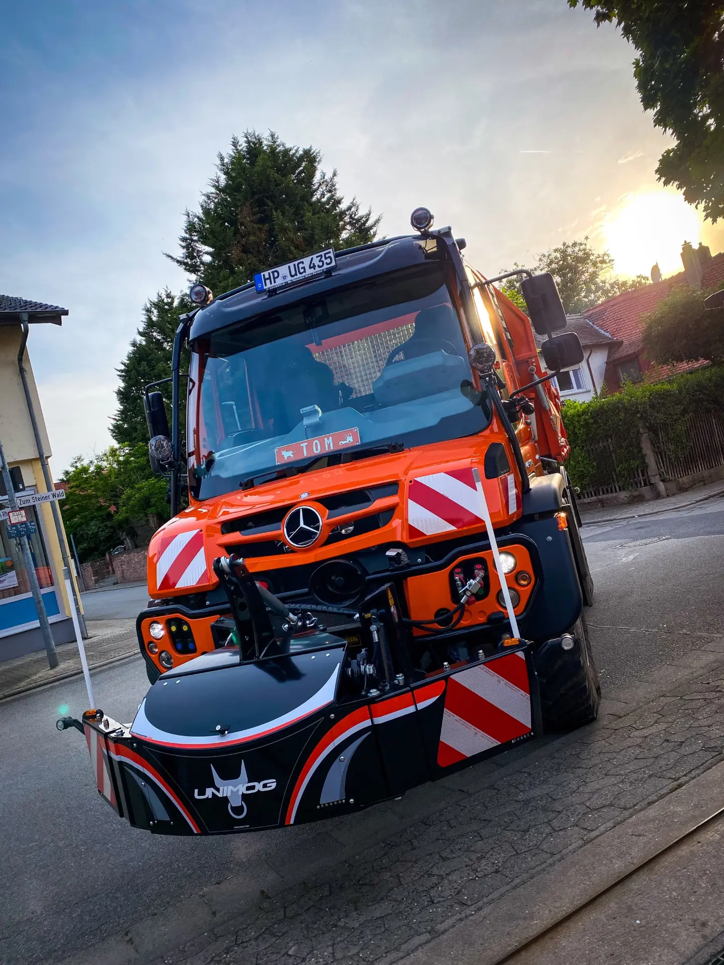 An orange Unimog with a black front weight bumper featuring red and white stripes and the Unimog logo
