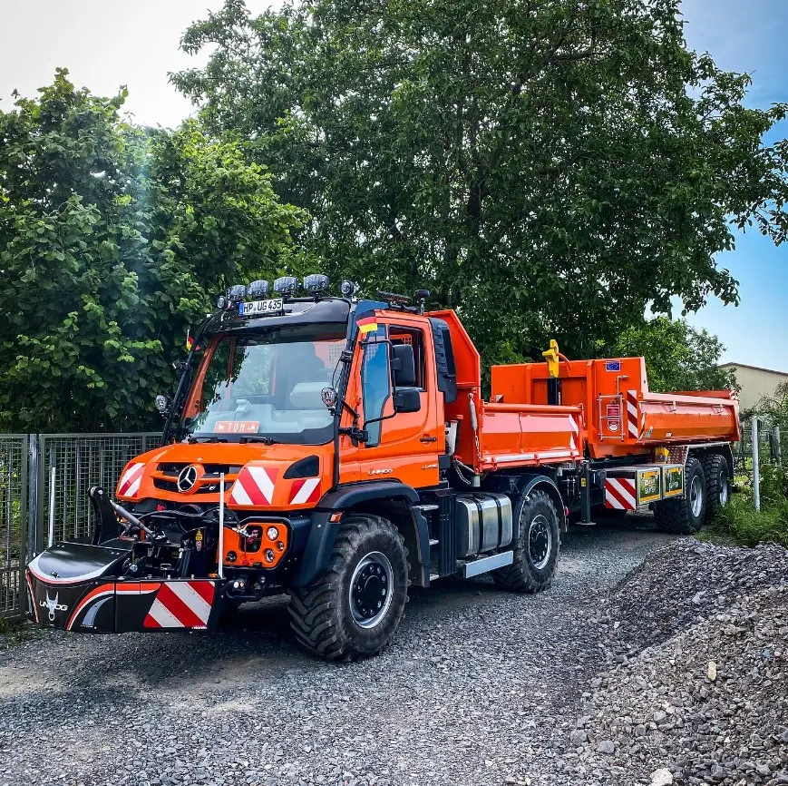 Orange utility vehicle with a front weight bumper, attached trailer, parked on a gravel path beside trees