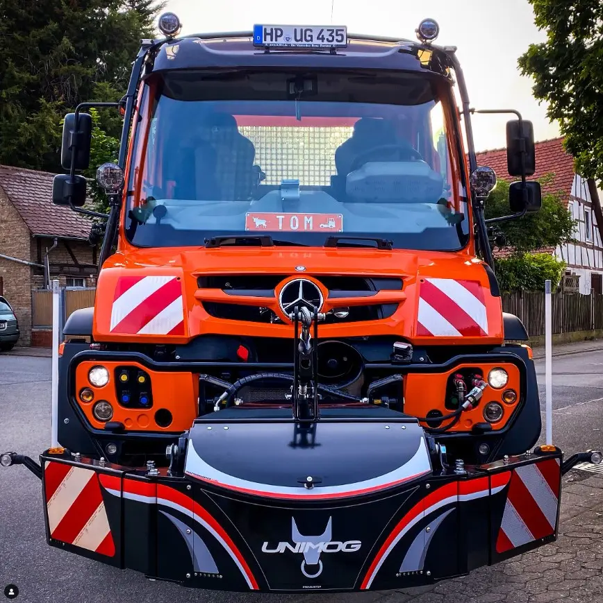 Orange Unimog vehicle with a large black and red front weight bumper featuring the UNIMOG logo and reflective stripes