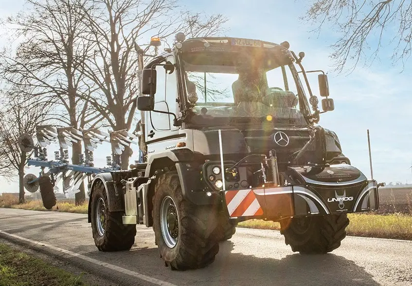 A tractor with a mounted front weight bumper labeled Unimog is parked on a rural road with farm equipment attached