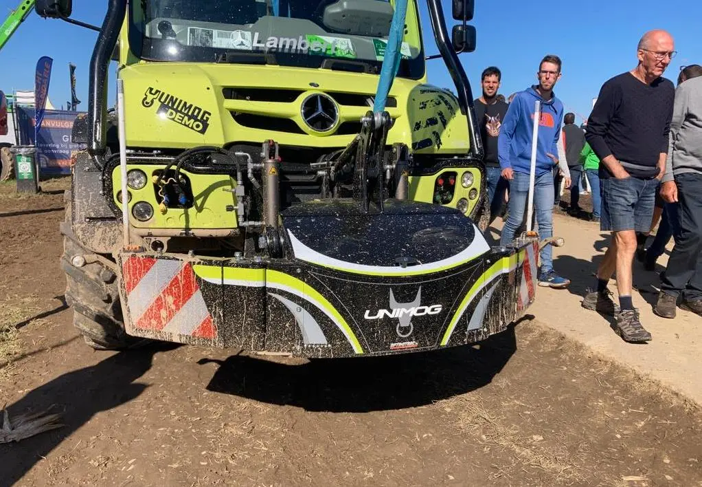 Muddy Unimog tractor with a front-mounted weight bumper, Unimog logo and safety stripes, surrounded by people