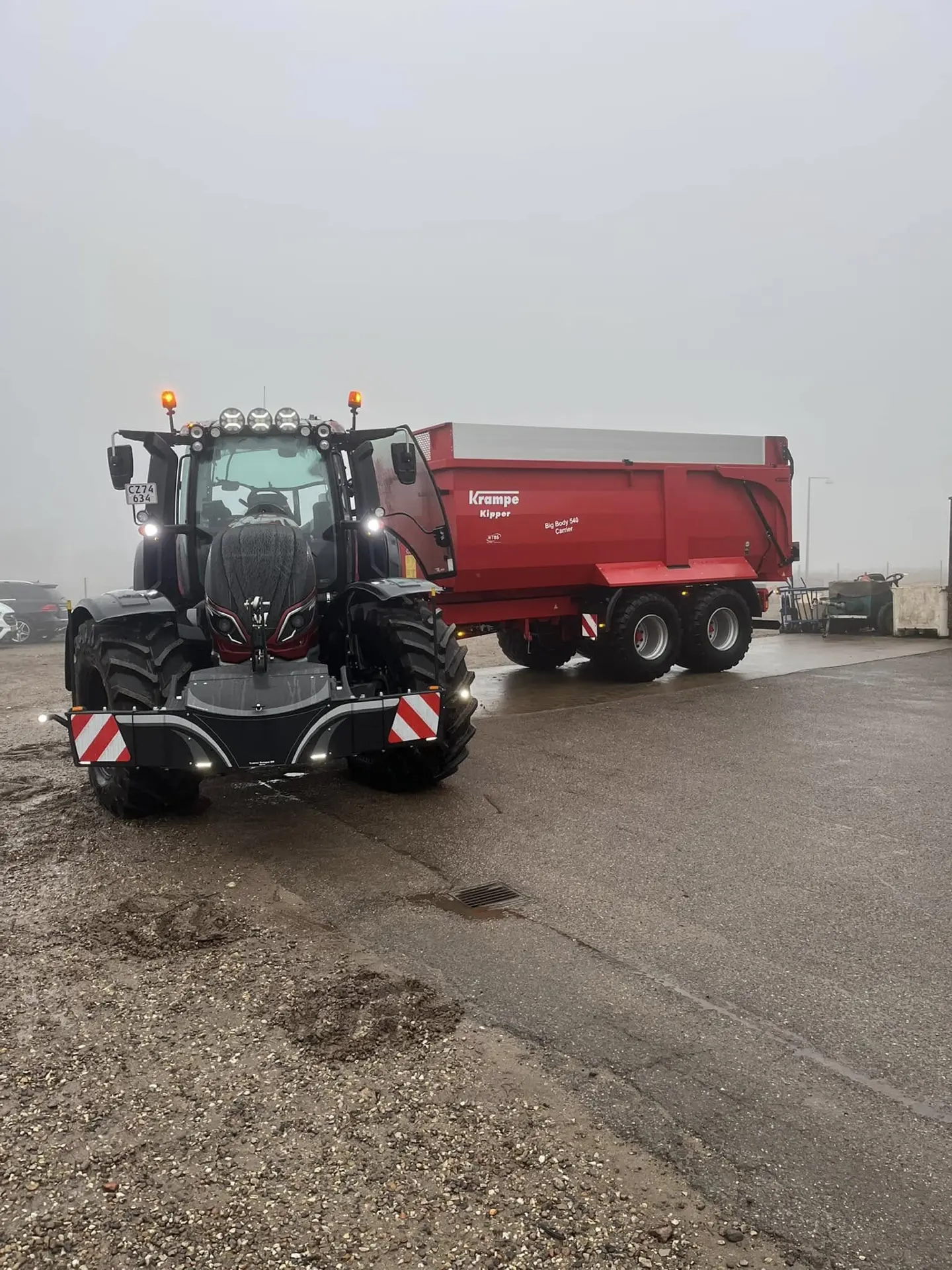 A tractor with a mounted front weight bumper and a red trailer attached, parked on a wet, overcast day