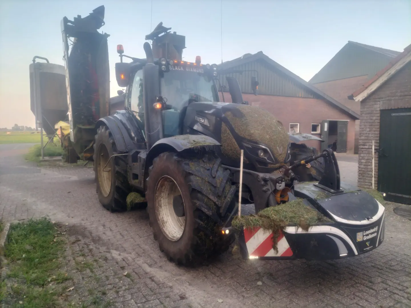 A tractor with grass-covered tires and a front weight bumper is parked on a farmyard near agricultural buildings