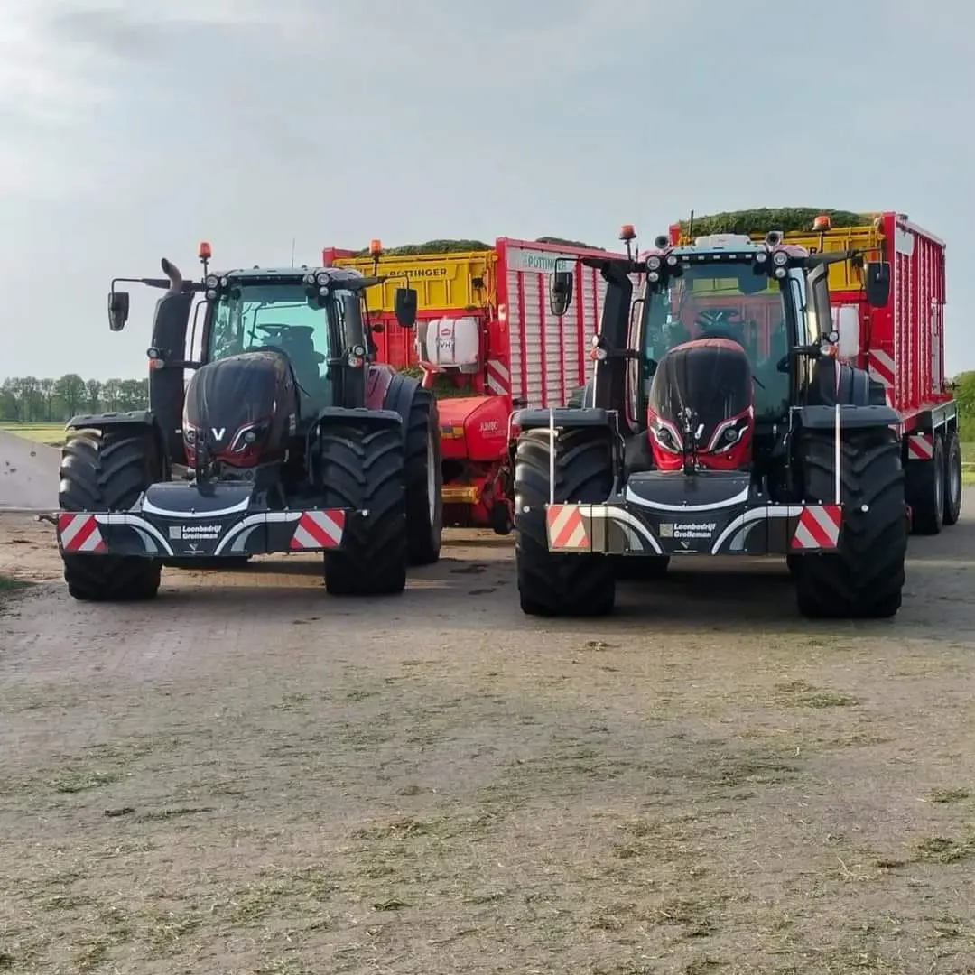 Two black tractors with front weight bumpers are parked side by side, each attached to a large red and yellow trailer
