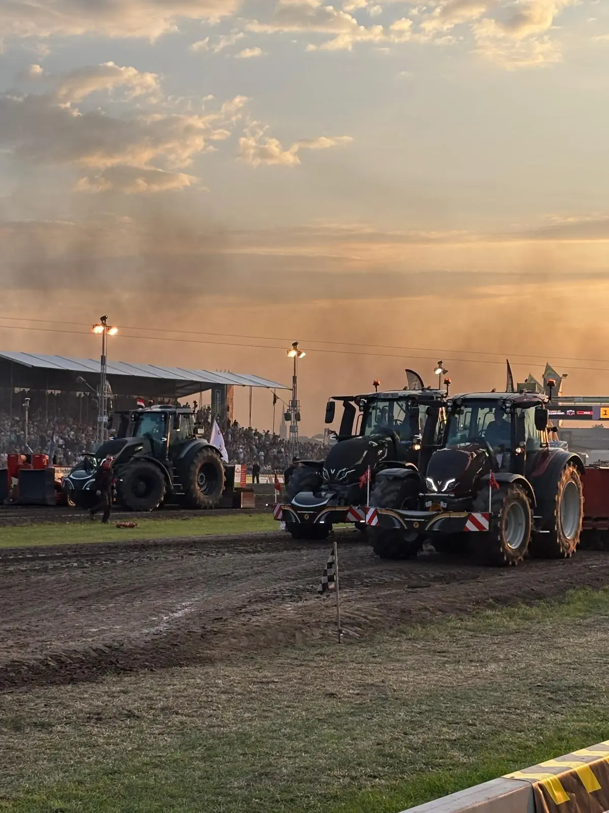 Three tractors with front weight bumpers are lined up on a dirt track at an outdoor event with a crowd in the background