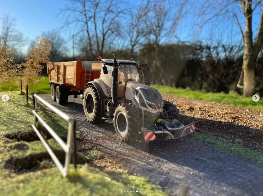 A tractor with a front weight bumper and attached trailer drives on a rural dirt road bordered by grass and trees