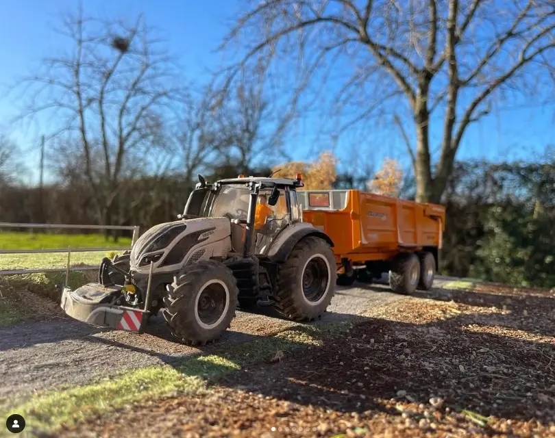 A tractor with a front weight bumper and an orange trailer is parked on a rural dirt path with trees in the background
