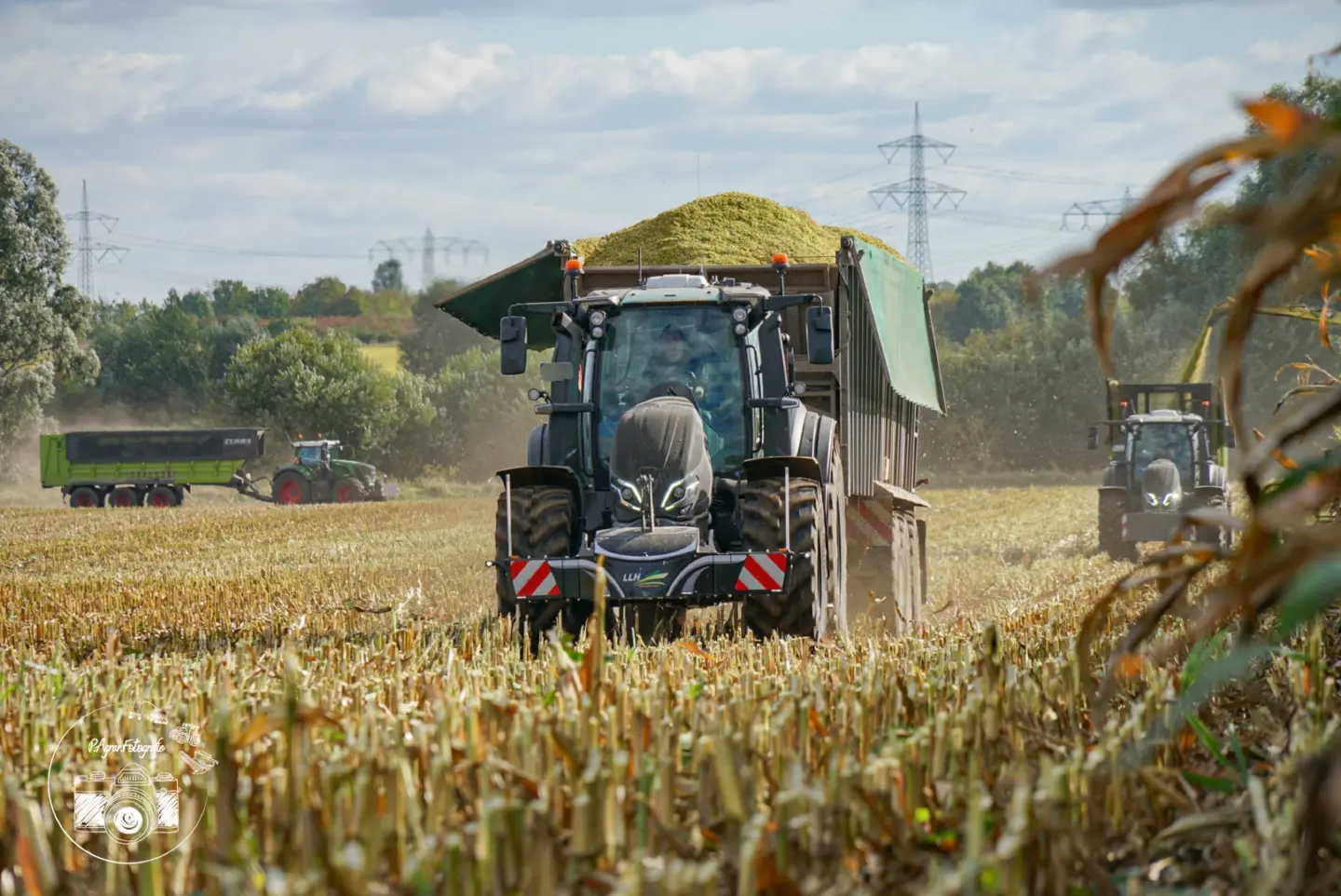 Black tractor with a front weight bumper towing a loaded trailer on a harvested field, two more tractors in background