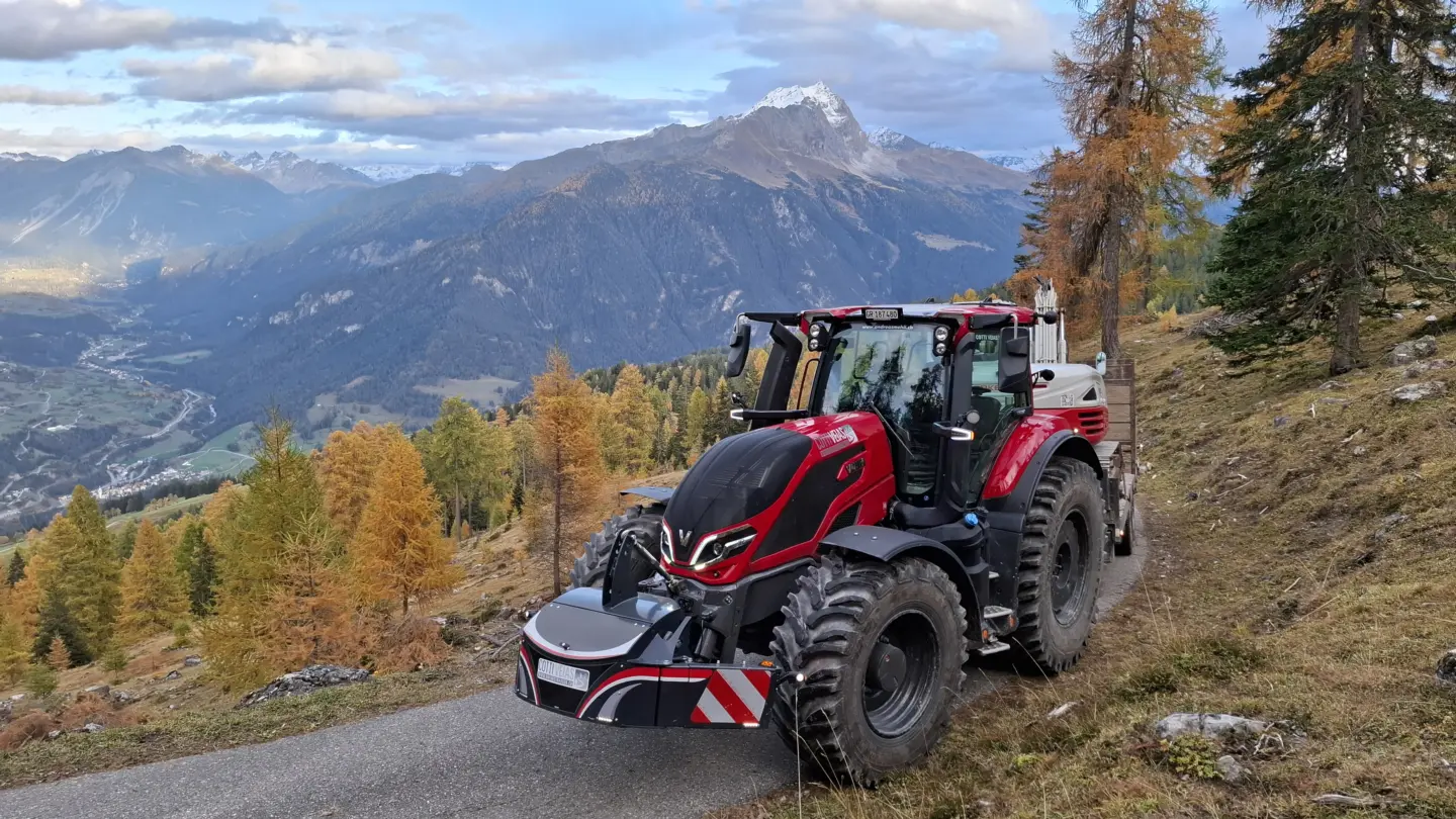 A red tractor with a front weight bumper attached is parked on a narrow mountain road with scenic valley views