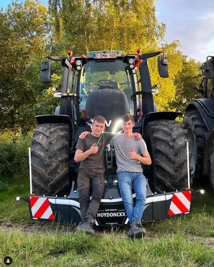 Two people sit on the front weight bumper of a large tractor parked on grass, both giving thumbs up gestures