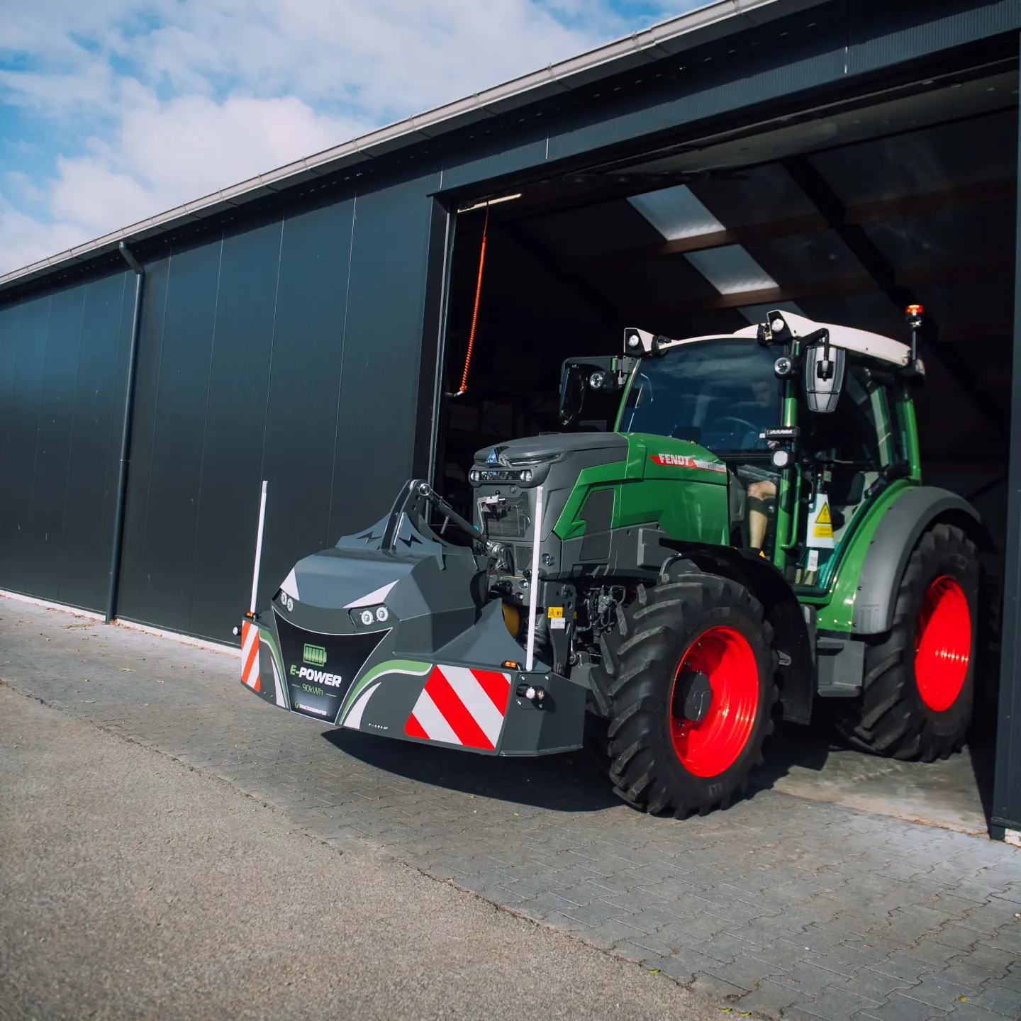 Green tractor with red wheels exiting a building, fitted with a large front weight bumper featuring warning stripes