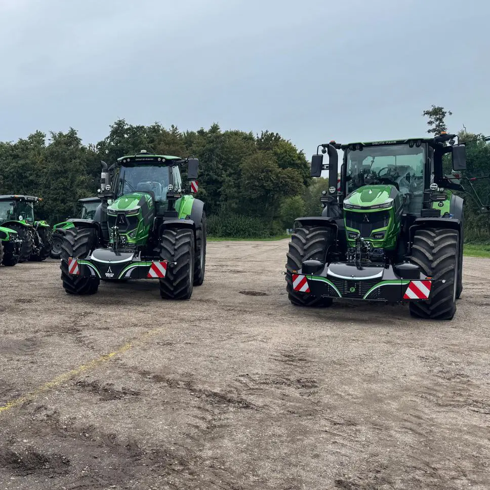 Two green tractors fitted with black front weight bumpers featuring red and white reflective panels on a dirt lot
