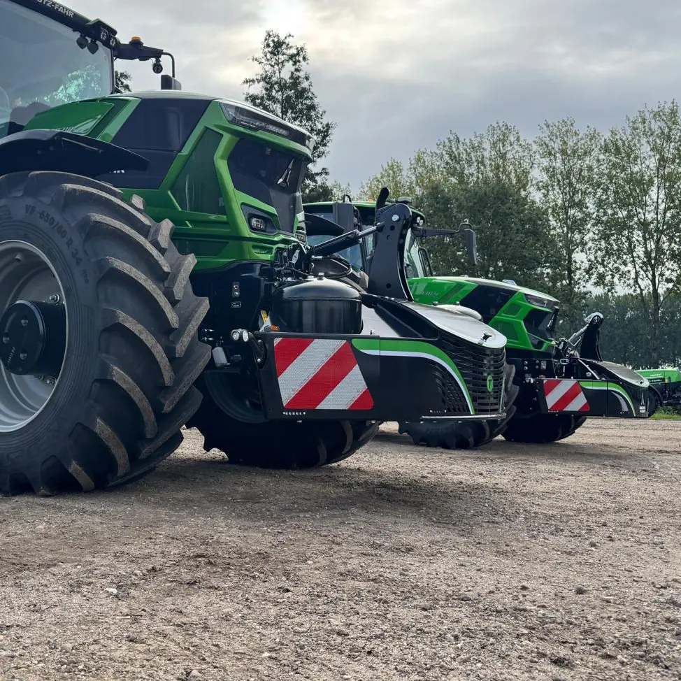 Two green tractors fitted with black front weight bumpers featuring red and white reflective stripes on a dirt lot